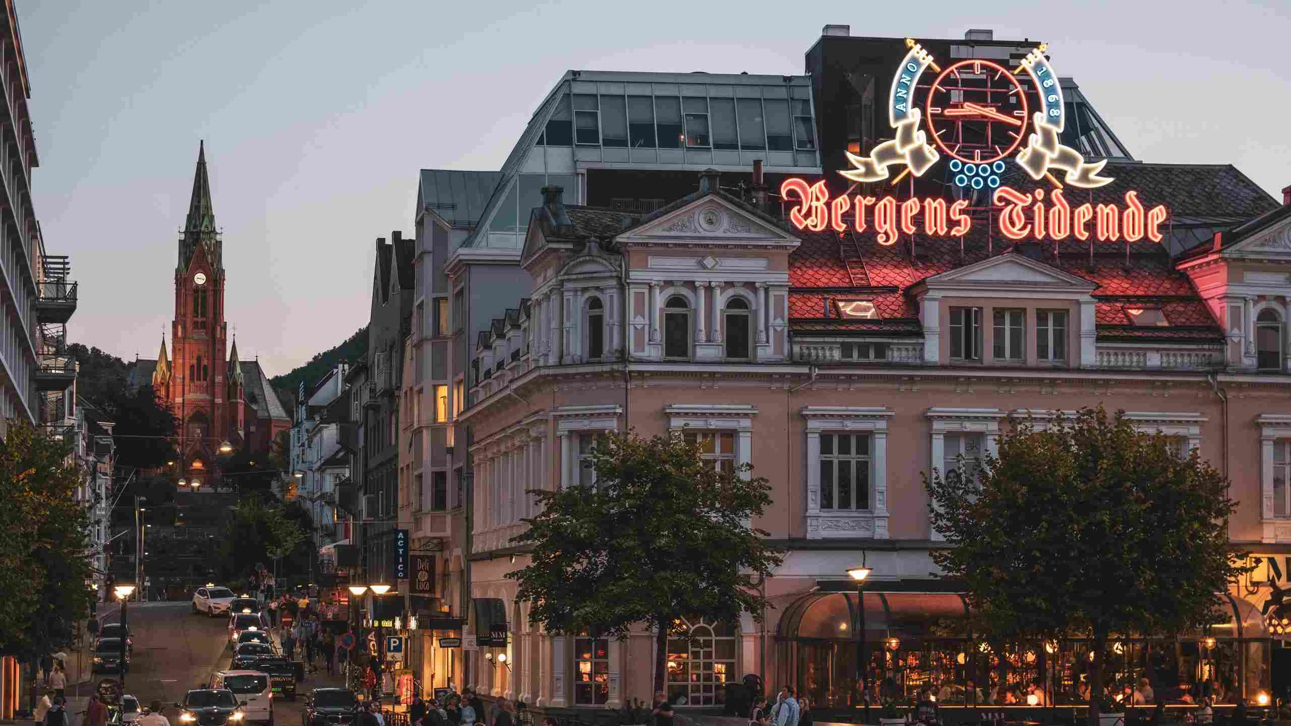 Image of a pink building with the "Bergens Tidende" logo on top. In front of the building there are people walking in different directions. Next to the building you can see a tall church. The picture was taken in the evening.