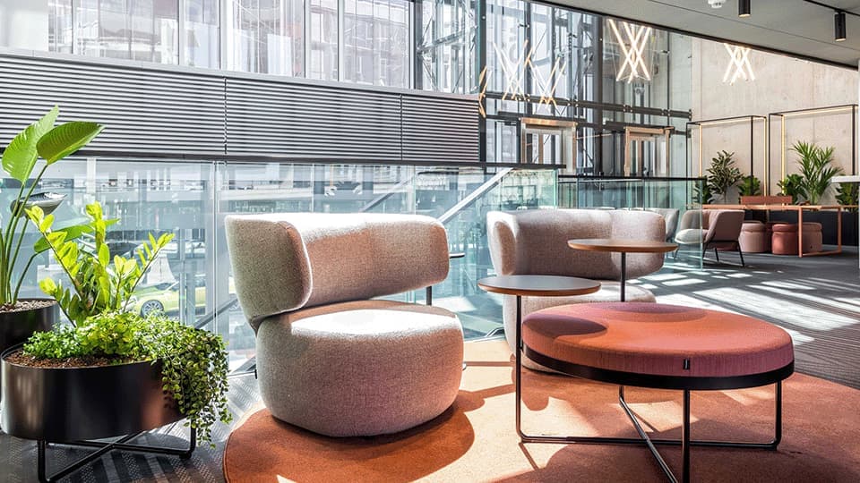 Photo of mezzanine in Tallinn lobby. Large windows with lots of daylight make the lobby seem even more open. In the picture, there are two large chairs in focus with a round table in front, placed on a large orange carpet. Two plants right next to the chairs.